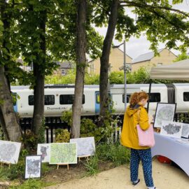 A woman in a yellow jacket peruses the Me on the Map stall. A Thameslink train is stopped at the platform in the background.