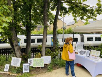 A woman in a yellow jacket peruses the Me on the Map stall. A Thameslink train is stopped at the platform in the background.